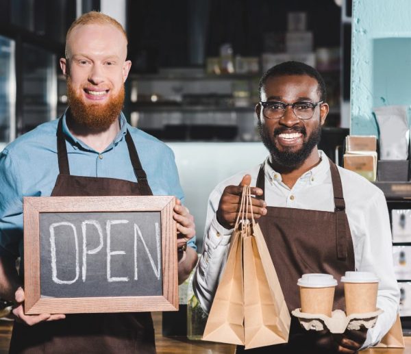 Small business owners in Sandy, UT smiling while holding coffee cups, paper bags and an open sign